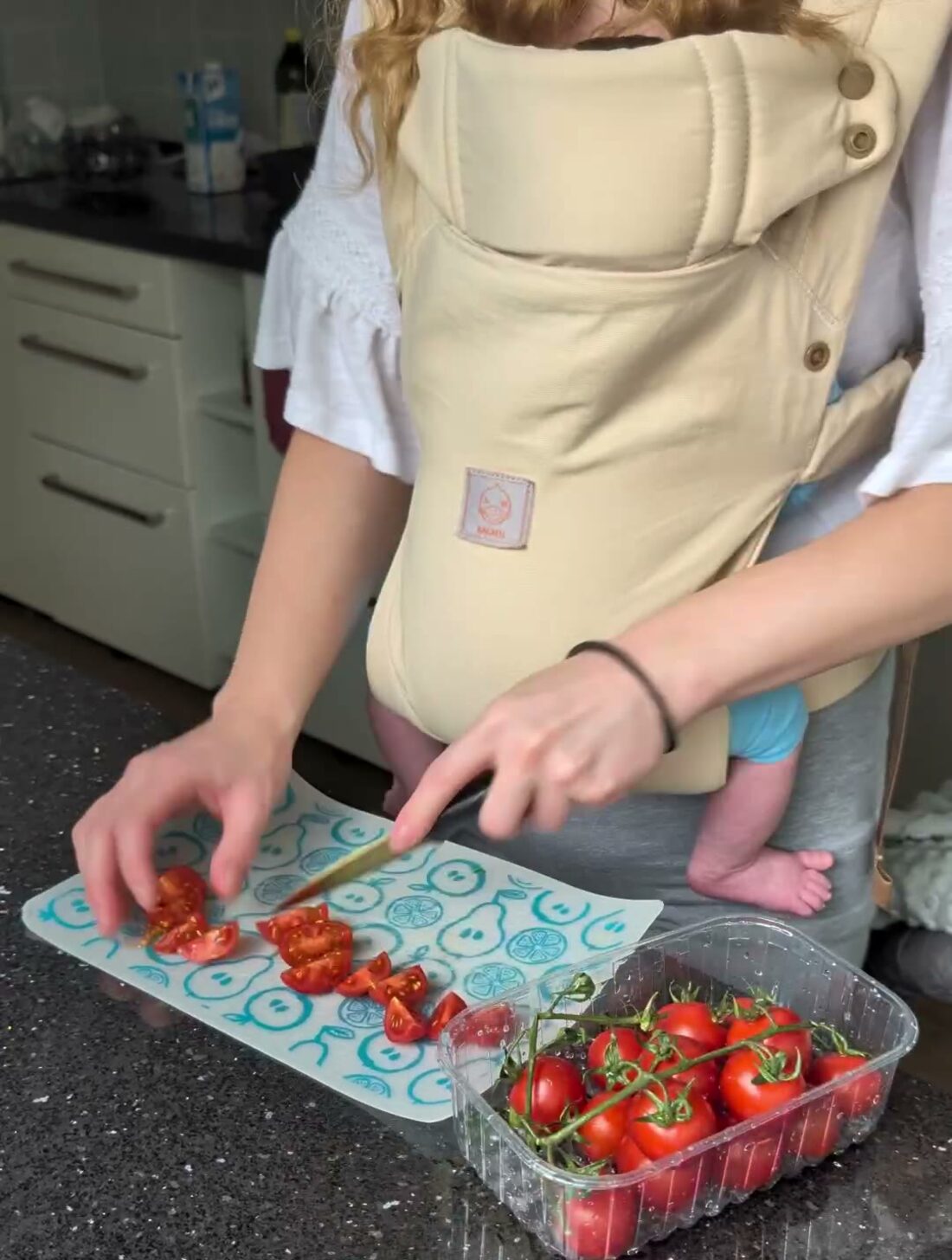 Hände schneiden Tomaten auf einem Schneidebrett; davor die Trage, in der das Baby sitzt.